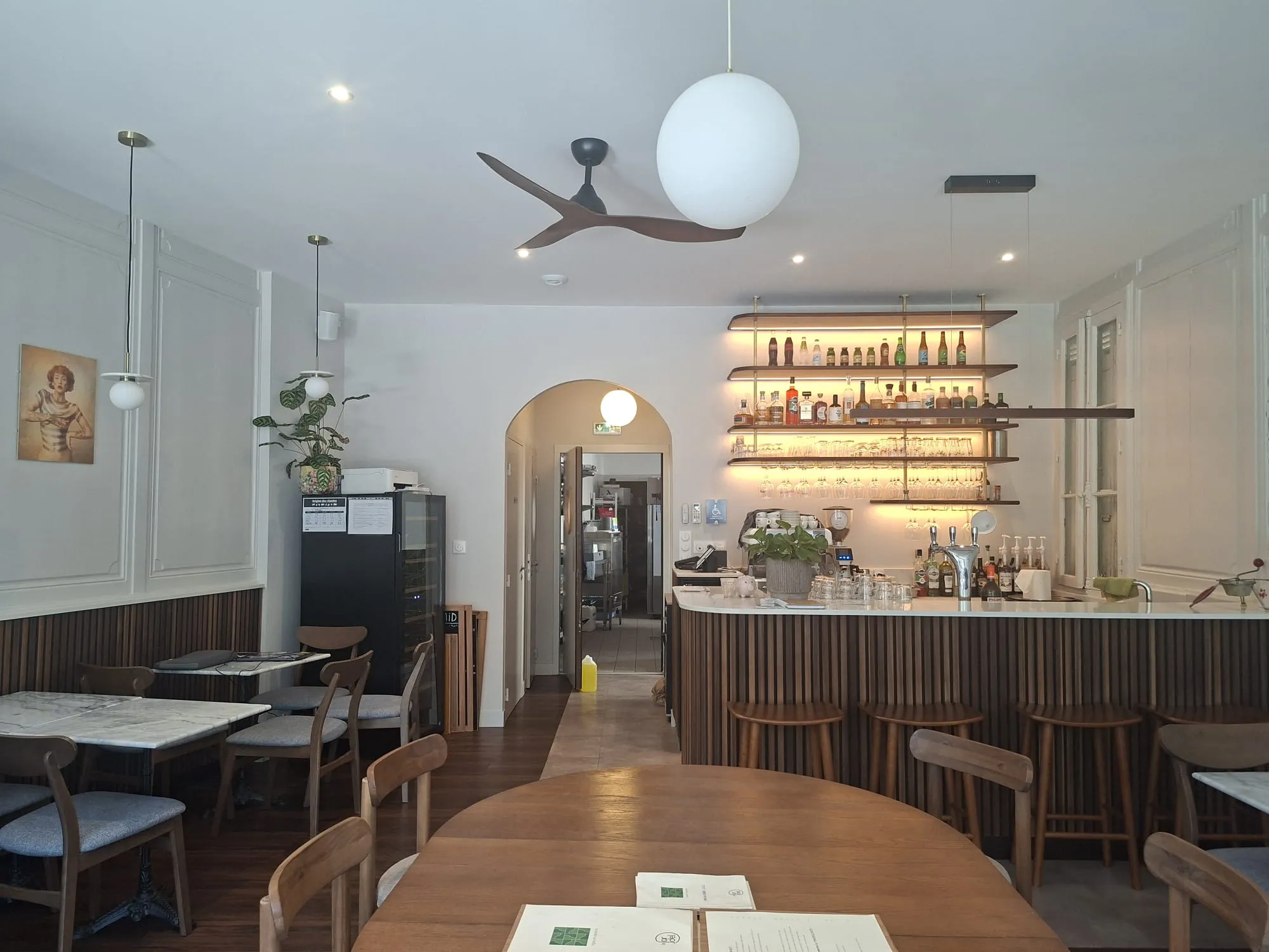 Salle du restaurant : comptoir en bois cannelé, étagères rétroéclairées, tables de marbre et moulures conservées